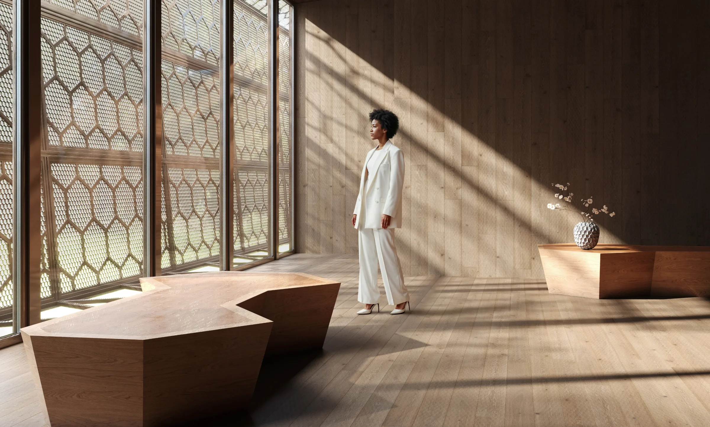 A woman in a light suit stands in a sunlit timber room beside sculptural furniture.