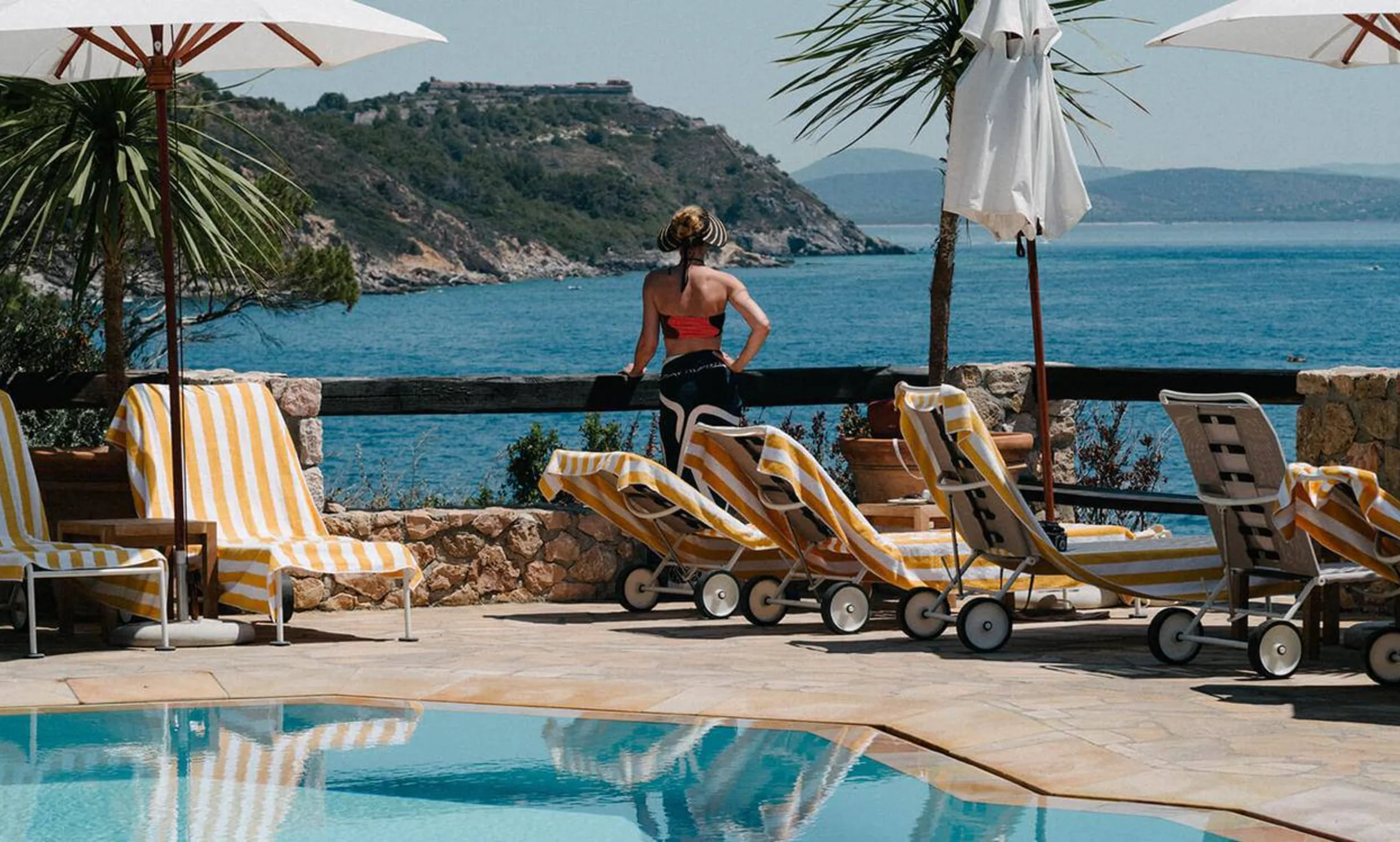A woman looks out over the sea beside a swimming pool and striped loungers.